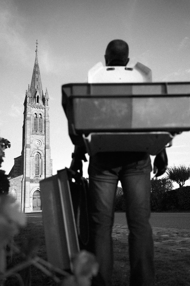 Clinet vineyard worker from behind holding wine crate looking up at Pomerol church