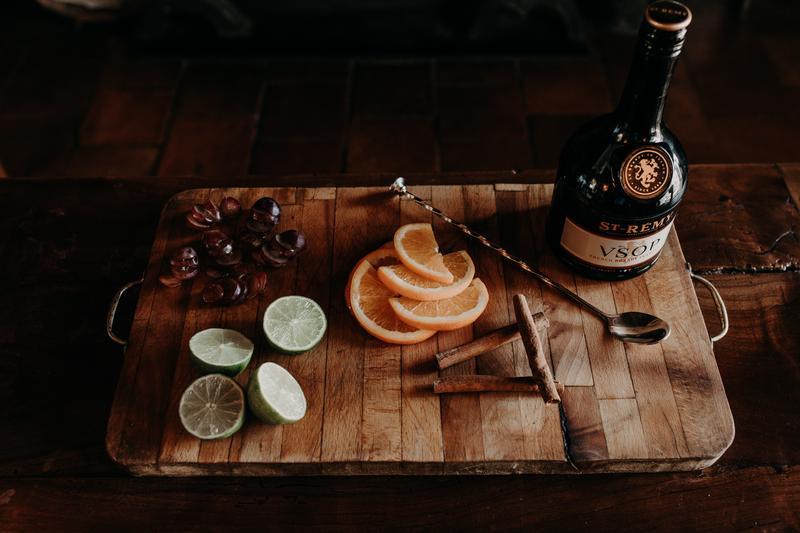 Top shot of chopping board lying on wooden table with fruit ingredients for cocktail and bottle of St-Rémy VSOP on top
