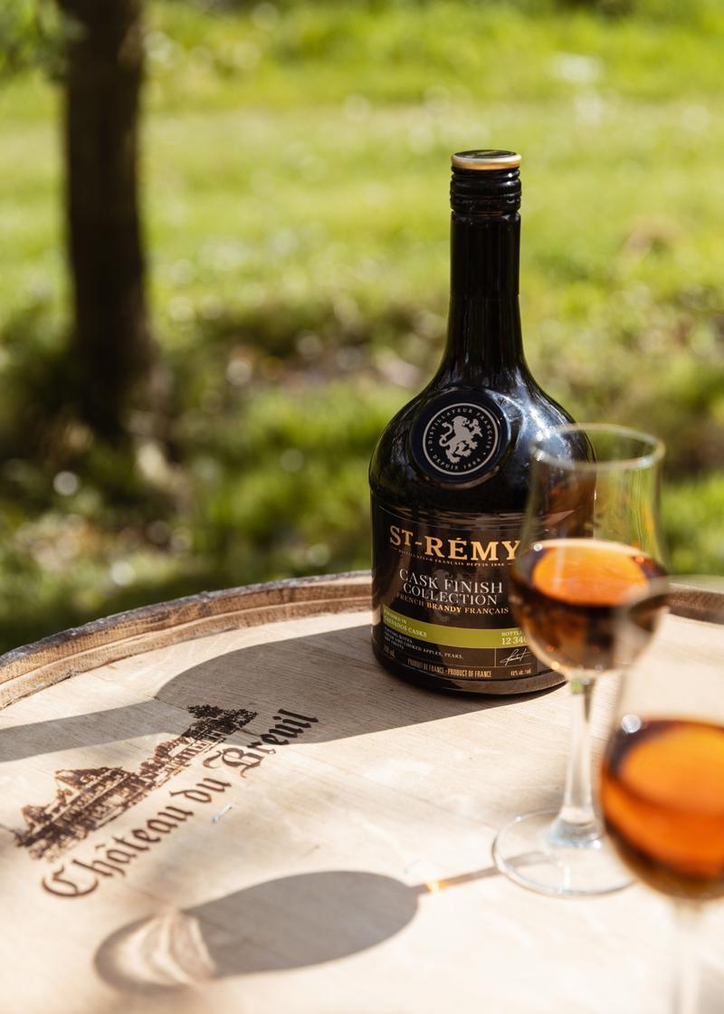 Bottle of St-Rémy calvados cask finish collection sitting on calvados barrel next to glass of brandy